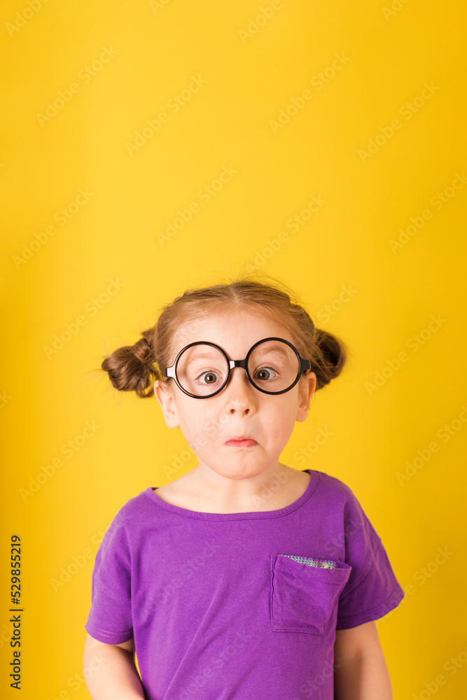 Little cute caucasian funny surprised kid child baby girl with glasses in purple T-shirt on yellow wall background. children studio portrait. Little cute caucasian funny surprised kid child baby girl with glasses in purple T-shirt on yellow wall background. children studio portrait.