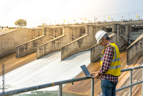 Aืn Engineer standing by the dam. He is wearing a white hard hat and yellow transparent vest.