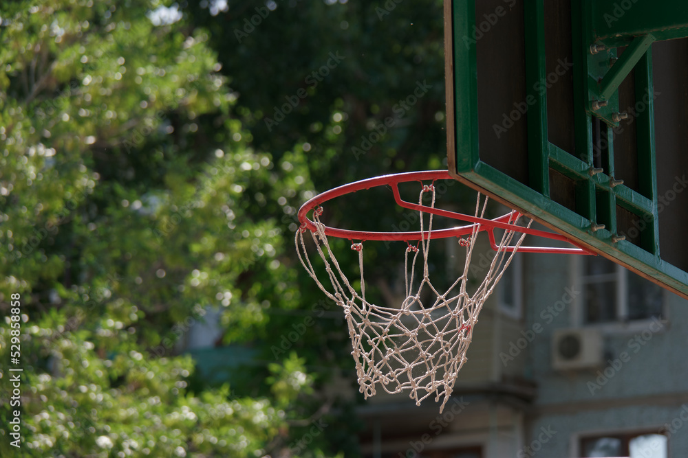 Rear view of red metal basketball hoop with white net and backboard against the green park trees and nearby building