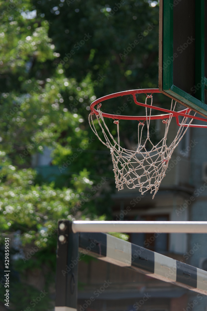 View of red metal basketball hoop with white net and backboard against the green park trees and nearby handball goal
