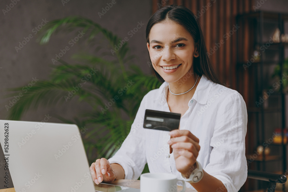 Young fun latin woman in white shirt use laptop pc computer credit bank card shopping online order delivery sit alone at table in coffee shop cafe restaurant indoor Freelance office business concept Young fun latin woman in white shirt use laptop pc computer credit bank card shopping online order delivery sit alone at table in coffee shop cafe restaurant indoor Freelance office business concept