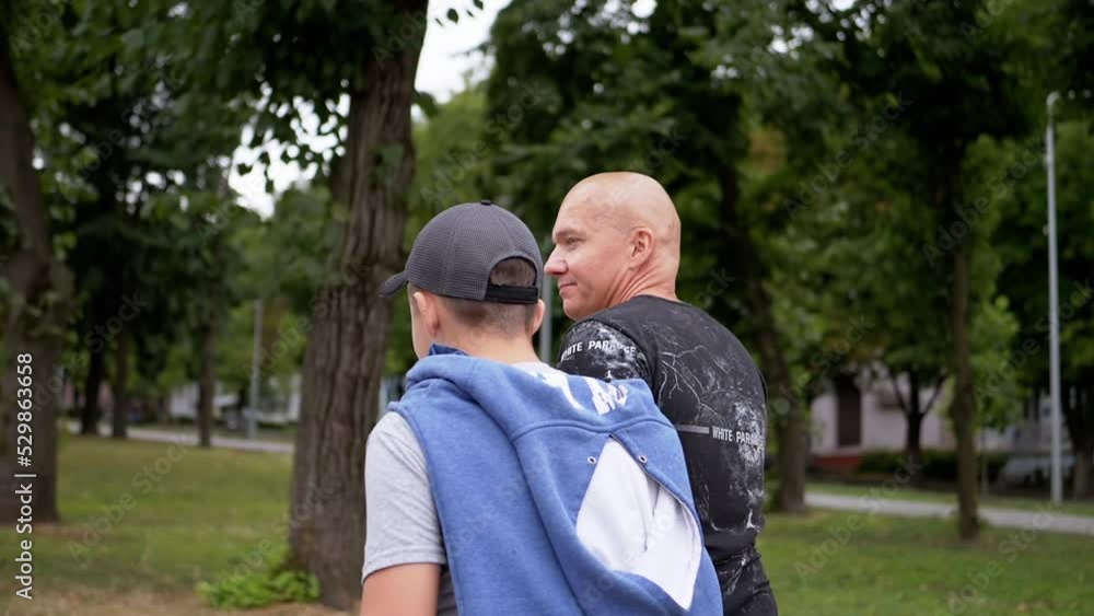 Back View Happy Father and Son Walking in the Park Outdoors in Cloudy Weather. A teenager in a hoodie and a cap is enjoying their vacation walking down the street with their bald dad. Recreation. Back View Happy Father and Son Walking in the Park Outdoors in Cloudy Weather. A teenager in a hoodie and a cap is enjoying their vacation walking down the street with their bald dad. Recreation.