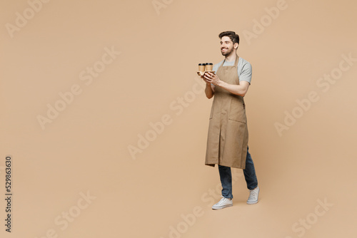 Full body side view young man barista barman employee in brown apron work in shop hold craft paper brown cup coffee to go walk isolated on plain pastel beige background Small business startup concept.