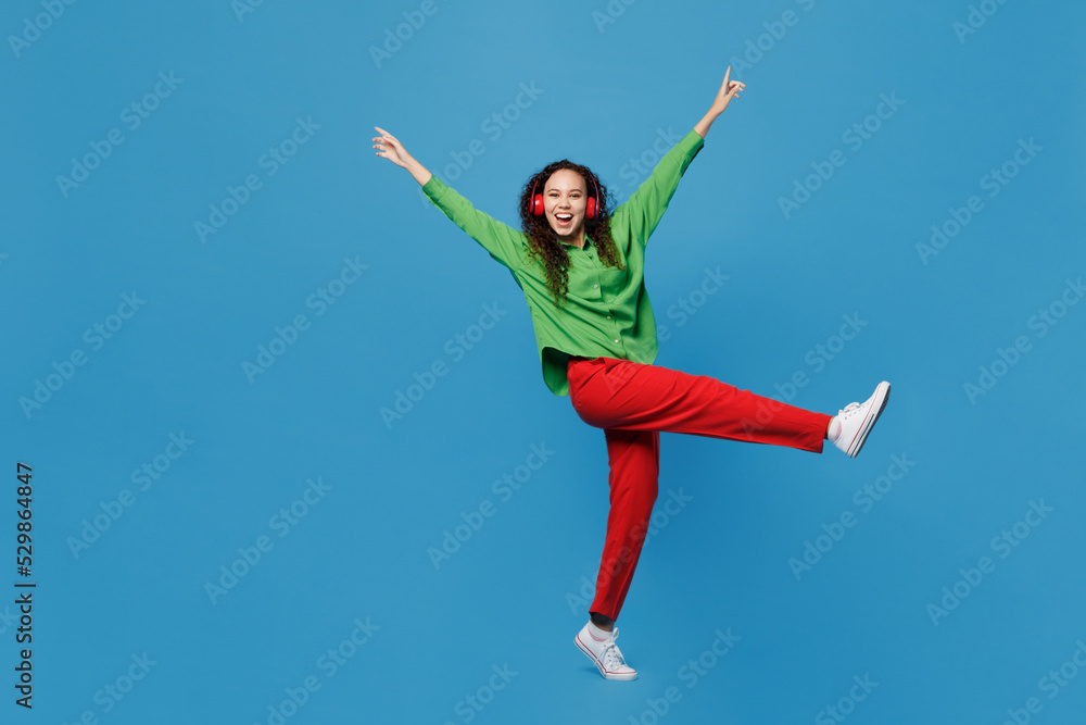 Full body side view young fun woman of African American ethnicity 20s she wear green shirt headphones listen music dance raise up hands legs isolated on plain blue background People lifestyle concept