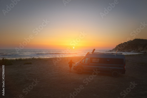 Silhouette of a campervan and woman on the beach against the orange setting sun. Adventure holiday, camping in the wilderness, nomadic life. Travelling in a van camper.