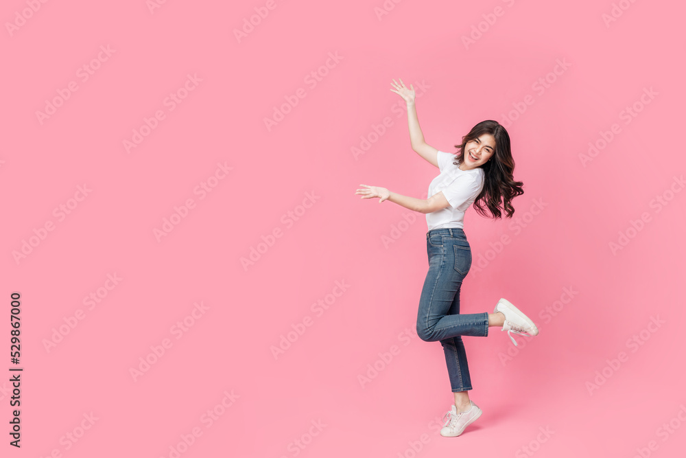 An asian woman in a casual outfit hopping off with her arm presenting, a cheerful expression on her face as she is excited to present something in isolated pink background.