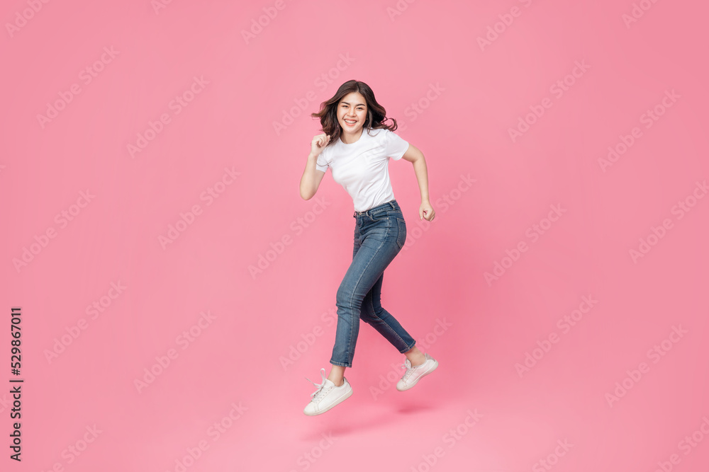Young Asian woman in a white t-shirt cheerful expression on her face as she is very happy over something excited smile and jumping with her arm raised in air on isolated pink background.