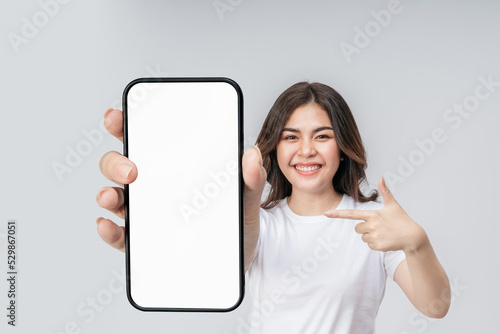 Selective focus, Smartphone blank screen with empty space for mobile app on screen on hand cheerful Asian woman. Smiling asian woman in white t-shirt pointing to the phone to recommending mobile app. © ME Image