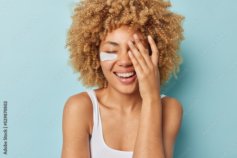 Positive curly haired woman covers face with palm giggles happily applies beauty patches under eyes to reduce wrinkles dressed in casual t shirt bare shoulders isolated over blue background. Positive curly haired woman covers face with palm giggles happily applies beauty patches under eyes to reduce wrinkles dressed in casual t shirt bare shoulders isolated over blue background.