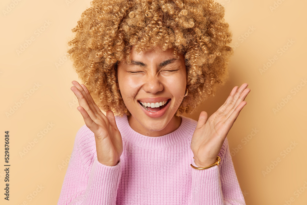 Overjoyed curly haired woman keeps palms raised laughs joyfully smiles broadly keeps eyes closed dressed in knitted pink jumper poses against beige background. Positive human emotions concept Overjoyed curly haired woman keeps palms raised laughs joyfully smiles broadly keeps eyes closed dressed in knitted pink jumper poses against beige background. Positive human emotions concept