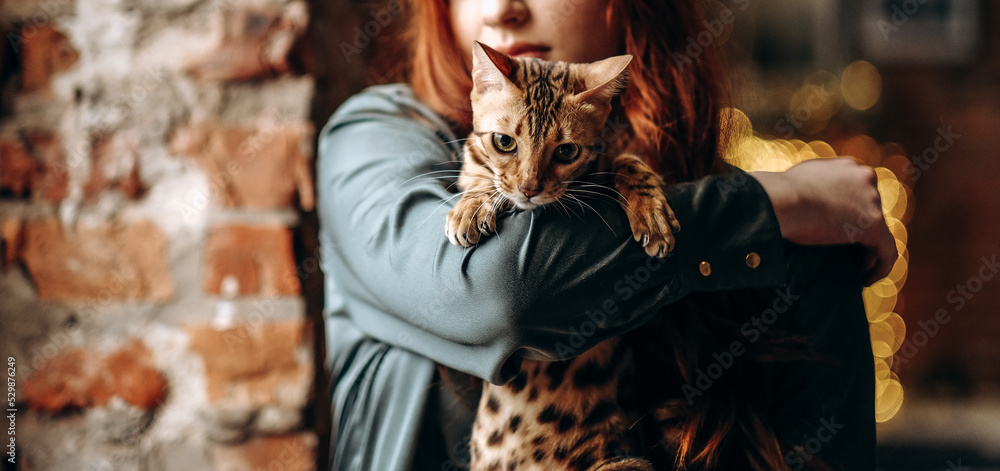 A young red-haired girl sits on the window and holds a Bengal cat.