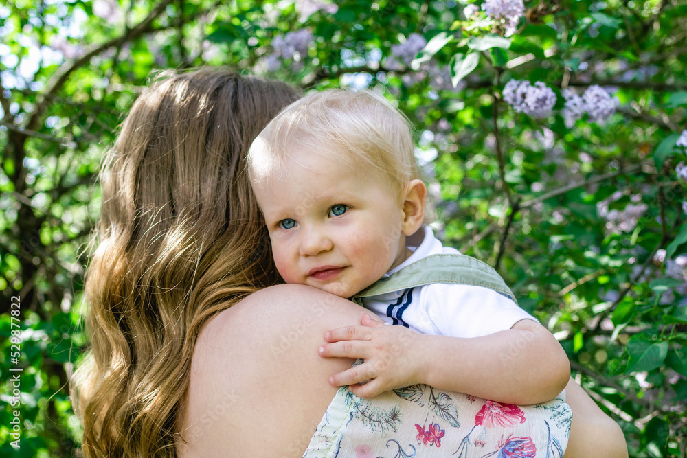 Portrait of mother with little son in her hands in park with lilac blossom tree, back view. Happy mother and child. Portrait of mother with little son in her hands in park with lilac blossom tree, back view. Happy mother and child.