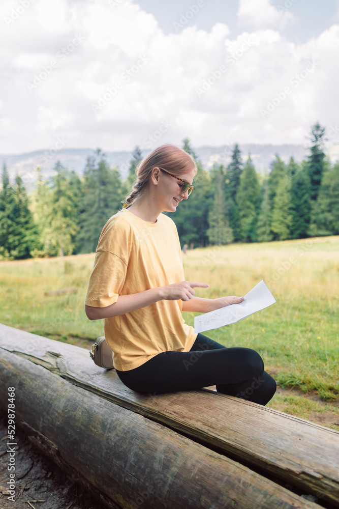 Young woman holding a map sits on the wood log while taking in the view from the top of a mountain. Girl tourist in mountain read the map. 