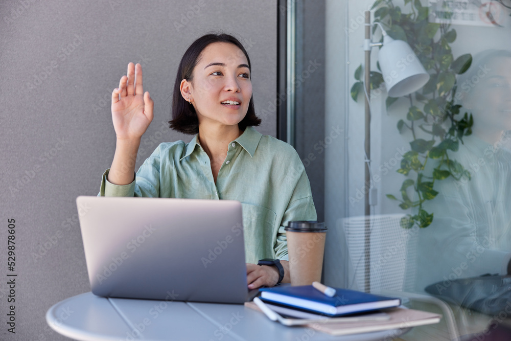 Pretty young Asian woman works distantly at laptop computer makes project waves hello someone poses at desktop with takeaway coffee and notepads uses modern technologies. Remote work concept Pretty young Asian woman works distantly at laptop computer makes project waves hello someone poses at desktop with takeaway coffee and notepads uses modern technologies. Remote work concept