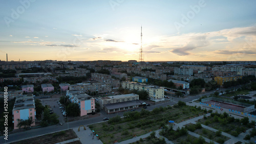 The bright sun behind the TV tower. The small town of Balkhash. There is one bird flying in the sky. Low houses, small streets and green trees. Cars are driving on the roads. Clouds in the sky