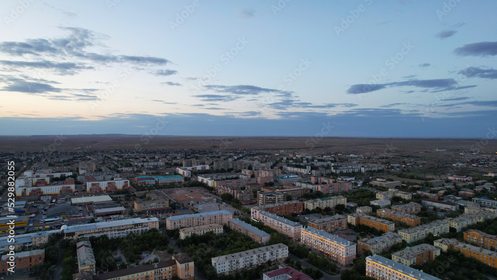 Dark clouds on the horizon. Sunset over a small town. Top view from a drone. Low colored houses are standing. Cars are driving on the road. Lights and house lights are on. The steppe is far away