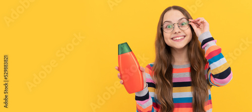 cheerful child long hair in glasses presenting shampoo bottle  advertisement. Banner of child girl hair care  studio poster header with copy space.