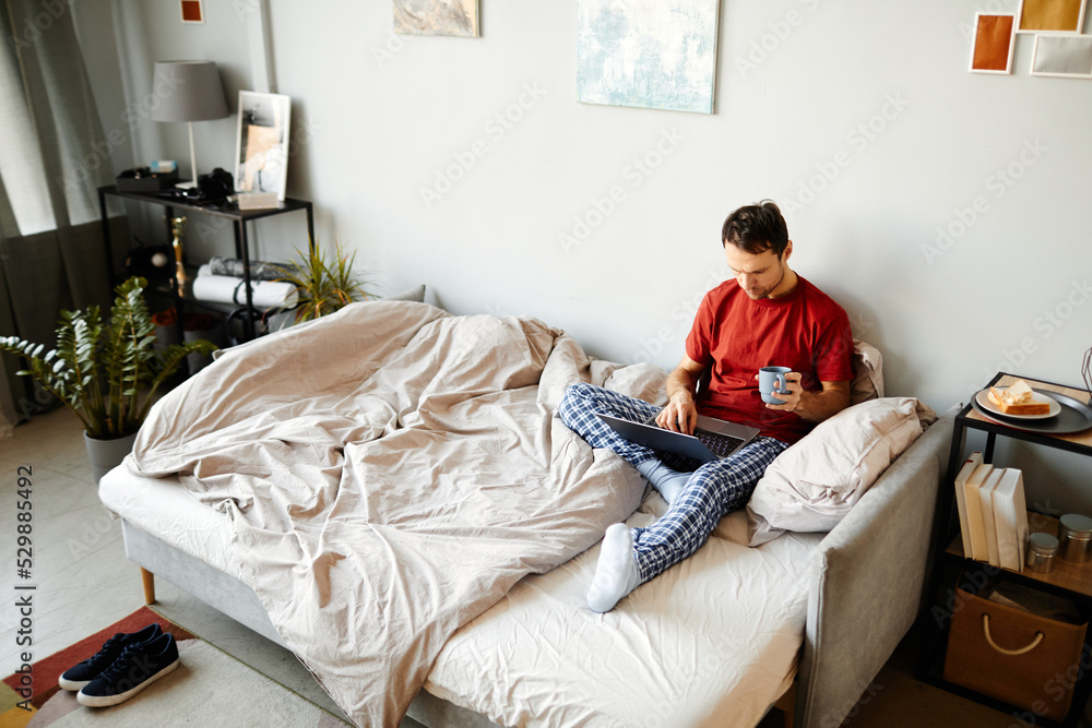 Obraz premium High angle view of young man sitting on bed and browsing in laptop with cup of coffee in the morning