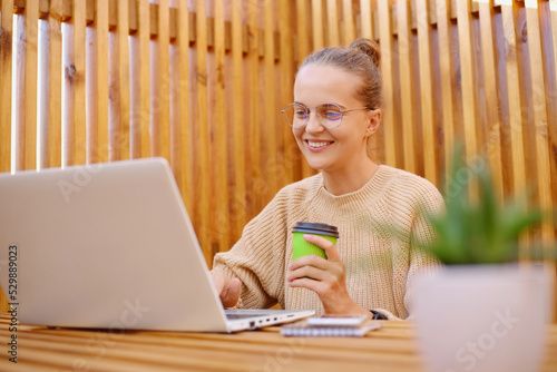 Portrait of smiling delighted satisfied woman freelancer with bun hairstyle wearing beige sweater working on laptop and drinking coffee, looking at display with happy expression.