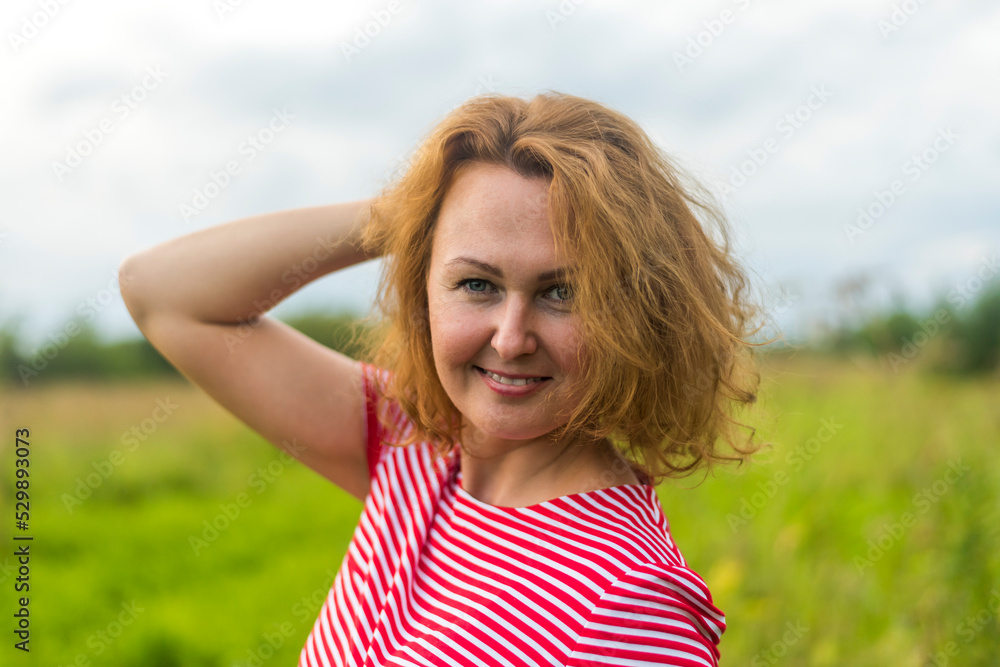 a girl with red hair in a red summer dress on the background of a green meadow and a sky with clouds a girl with red hair in a red summer dress on the background of a green meadow and a sky with clouds
