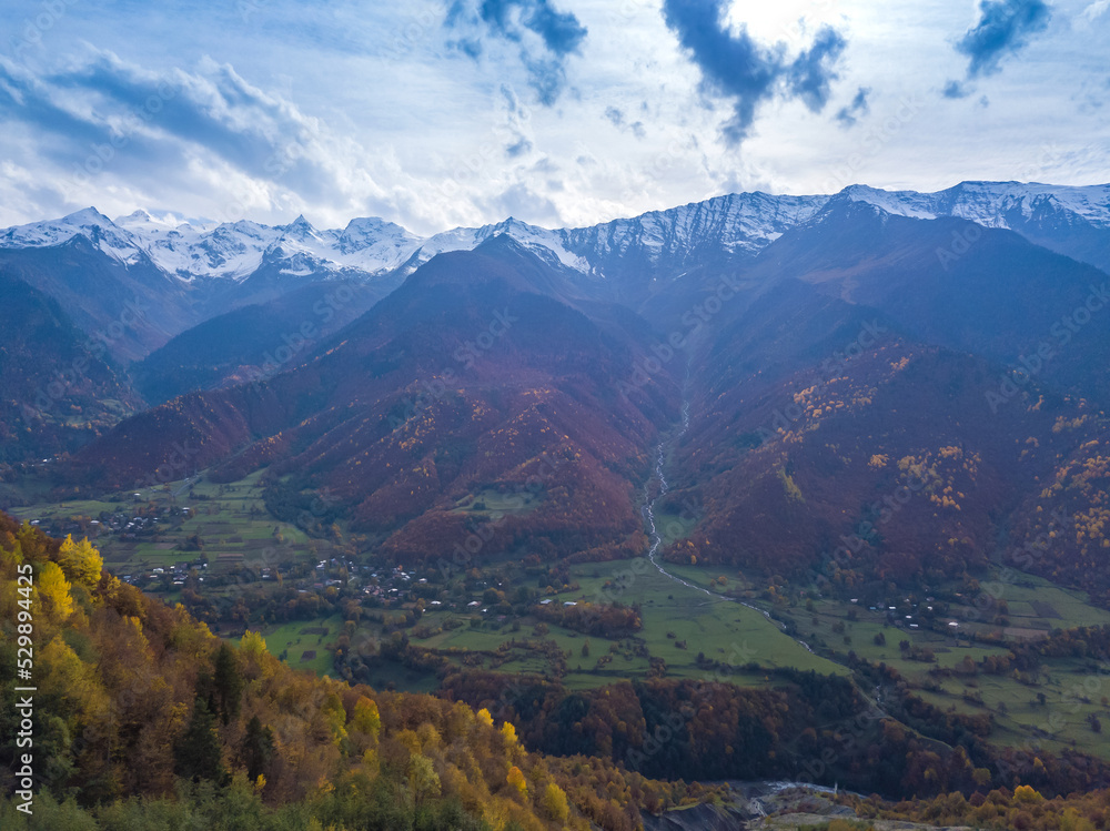 Beautiful view from a drone of a village at the foot of the mountains on an autumn day. Beautiful autumn landscape with autumn trees, snow-capped mountains