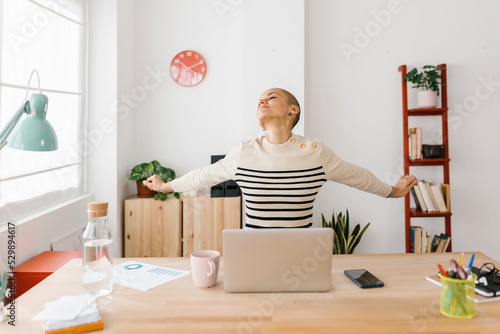 Smiling mid adult woman stretching body while working on laptop at home office - Technology, remote work and business concept © Xavier Lorenzo