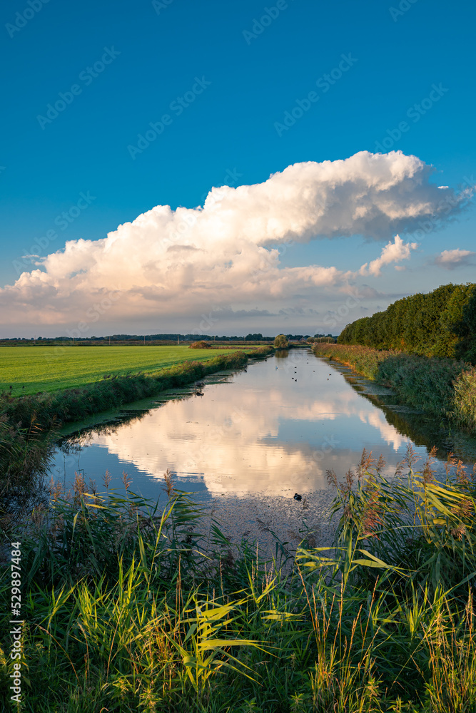 Dissipating storm cloud is reflected in the calm water of a canal on a serene summer evening