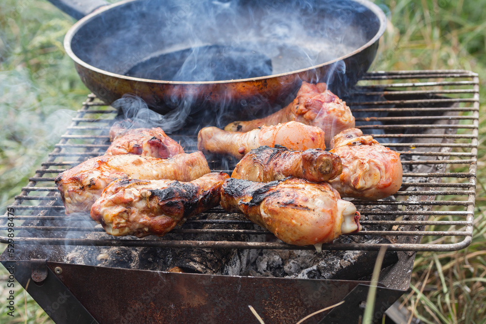 chicken drumsticks on a grill next to a frying pan in nature, side view