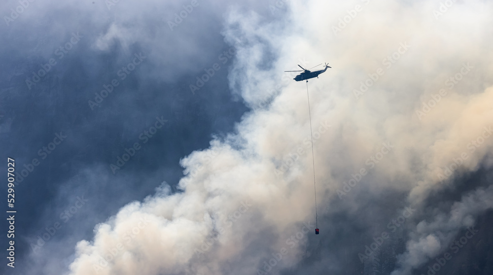 Wildfire Service Helicopter flying over BC Forest Fire and Smoke on the mountain near Hope during a hot sunny summer day. British Columbia, Canada. Natural Disaster Wildfire Service Helicopter flying over BC Forest Fire and Smoke on the mountain near Hope during a hot sunny summer day. British Columbia, Canada. Natural Disaster