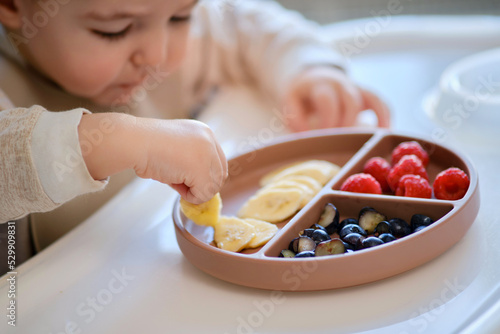 Toddler baby eats fruits and berries with his hand, table close-up. Child hands take food from a beige plate. Kid aged one year and two months