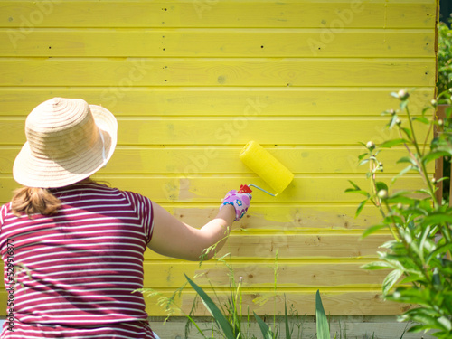 young girl paints a wooden wall with a roller with yellow paint