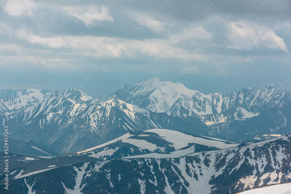 Fototapeta premium Awesome gloomy aerial view to high snow mountain peak in overcast. Atmospheric mountain landscape at high altitude with cloudiness. Alpine scenery with snowy mountain top in center under cloudy sky.