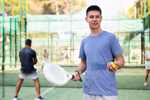 Positive confident sporty young guy standing on paddle tennis outdoor court on summer day with racket and ball in hands, ready for friendly match on blurred background with players © JackF