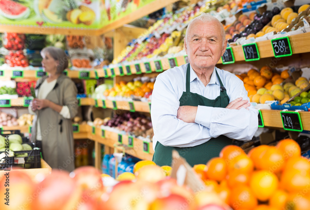 Old man supermarket worker standing in salesroom amongst shelves with vegetables and fruits and looking in camera. Woman making purchases in background.