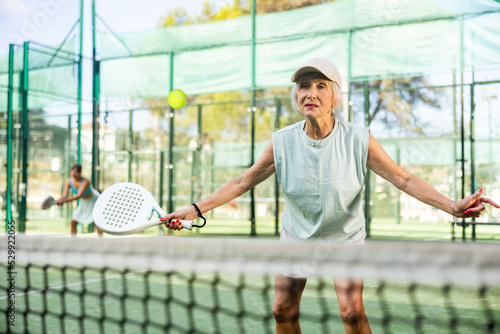 Sporty mature woman padel player hitting ball with a racket on a hard court © JackF