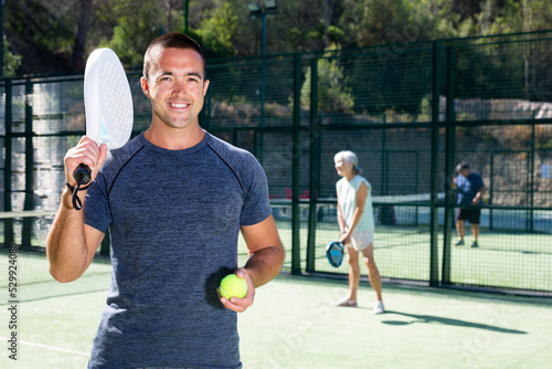 Smiling sporty man standing on outdoor paddle tennis court with racket and ball in hands, ready for friendly match on blurred background with people playing on summer day © JackF