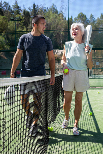 Sporty woman and man in sportswear walking on outdoor court along net with paddle racket and ball in hands after match, talking friendly © JackF