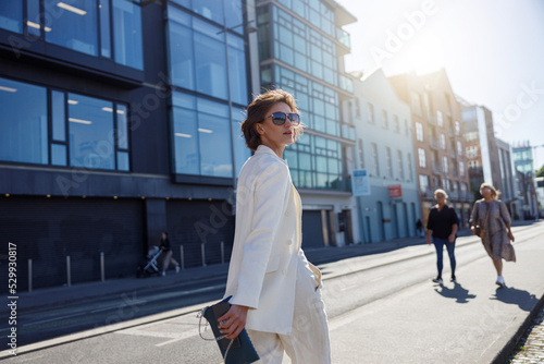 Woman in trendy outfits crosses road against backdrop of city buildings. Blurred background