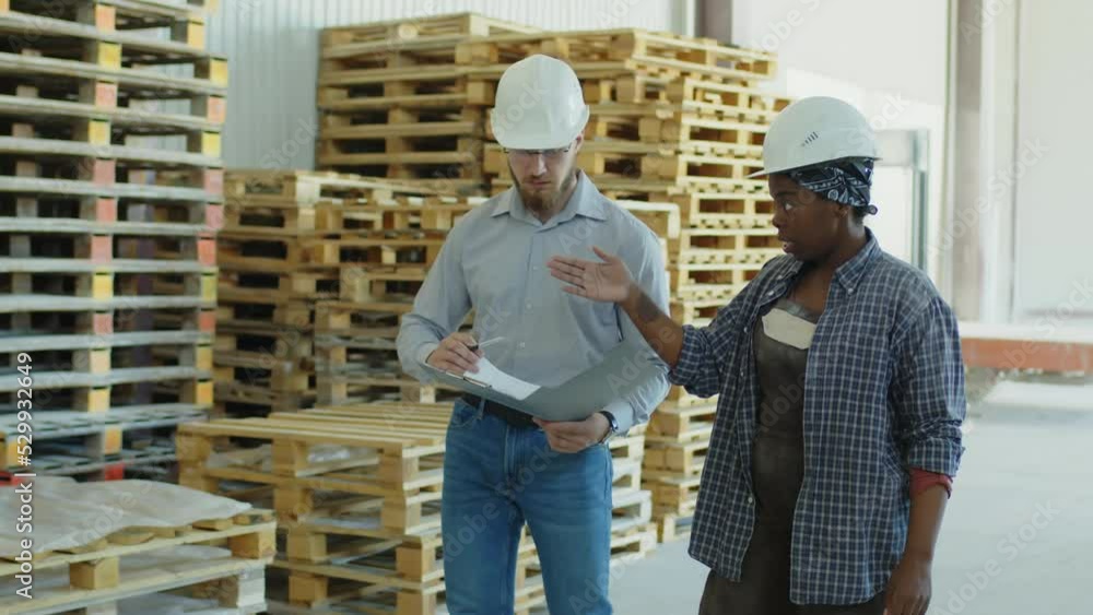 Medium shot of foreman talking to African American female employee while walking along wooden pallets at warehouse Medium shot of foreman talking to African American female employee while walking along wooden pallets at warehouse