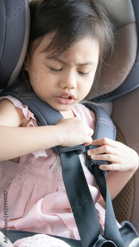 Dramatic portrait of 4 years old cute baby Asian girl, little preschooler child fastened with security seat belts, sitting in child seat in car. Kid try to unbuckled car seat belt photo