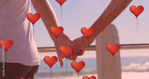 Multiple heart balloons floating against mid section of couple holding hands at beach