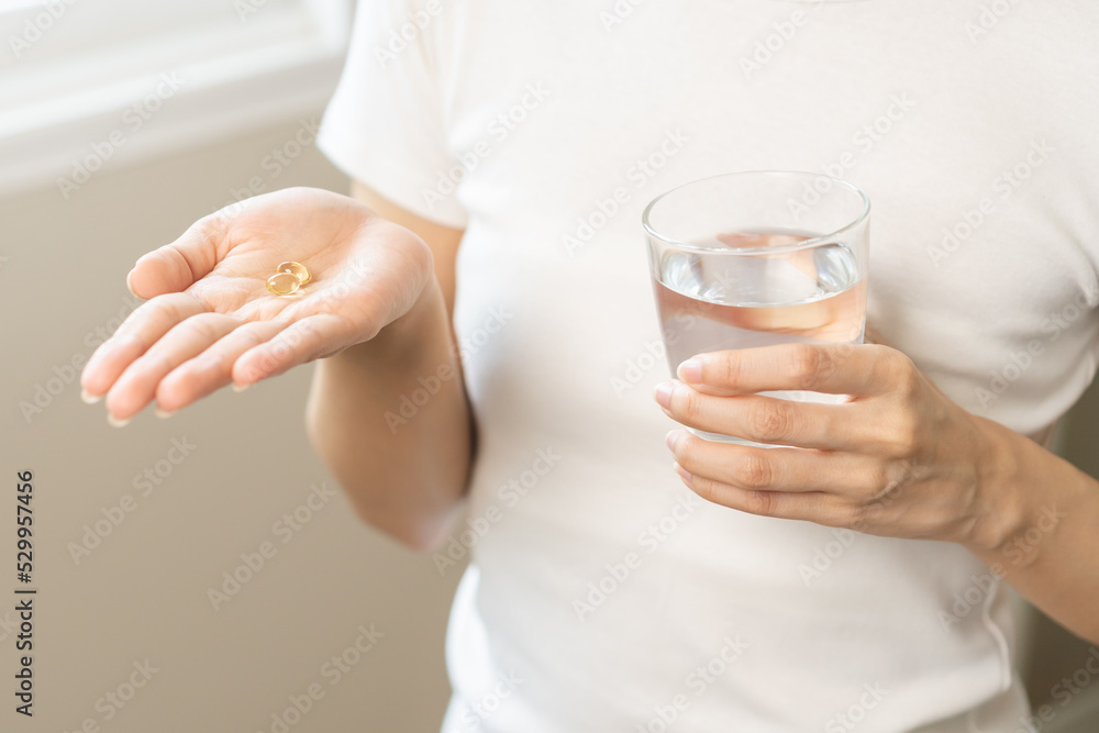 Dietary supplement, asian young woman hand holding fish oil or medical pill, take or eat vitamin C, D for treatment for skin, hair and nail strengthen with glass of pure water at home, healthcare. Dietary supplement, asian young woman hand holding fish oil or medical pill, take or eat vitamin C, D for treatment for skin, hair and nail strengthen with glass of pure water at home, healthcare.