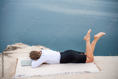 A woman is lying and typing on a laptop keyboard on a terrace with a beautiful sea view. Wearing a white blouse and black skirt. Freelance travel and vacation concept, digital nomad.