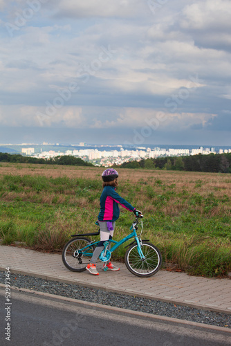 Fototapeta Naklejka Na Ścianę i Meble -  A little girl on her bicycle looks at the landscape. A child on a bicycle is stopped and looks at the landscape in front of her. In the distance is a large city.