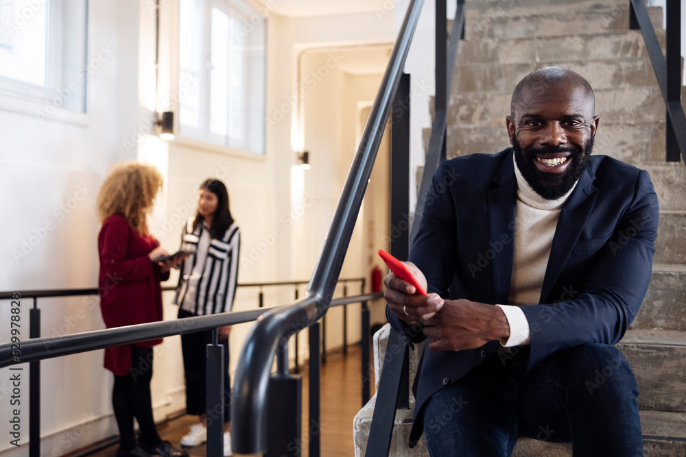 Smiling businessman with smartphone sitting on stairs colleagues talking in background Smiling businessman with smartphone sitting on stairs colleagues talking in background