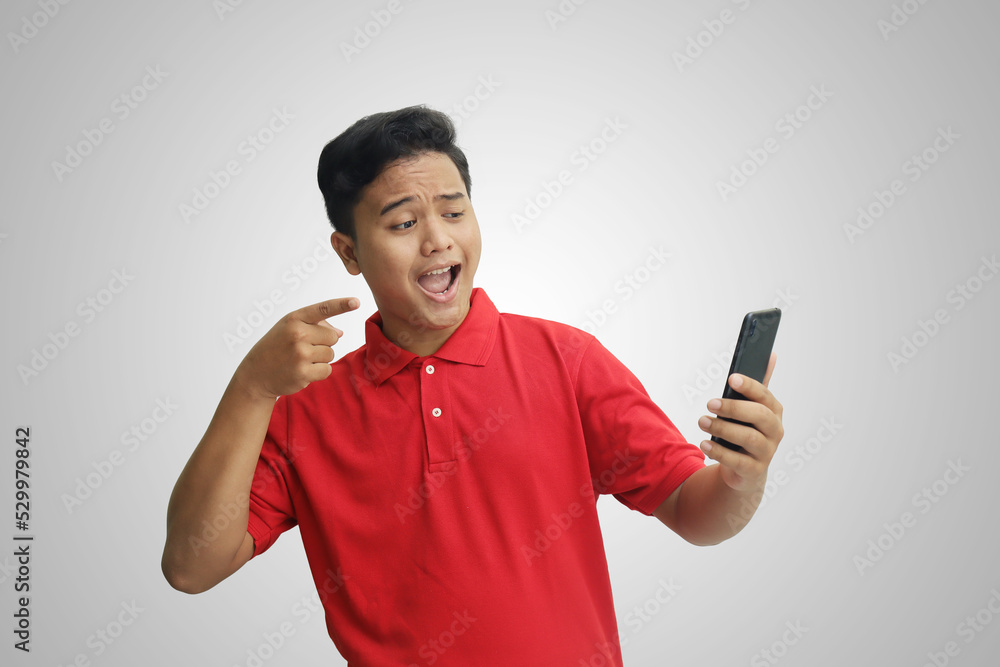 Portrait of cheerful Asian man in red polo shirt pointing his index finger at mobile phone. Isolated image on gray background Portrait of cheerful Asian man in red polo shirt pointing his index finger at mobile phone. Isolated image on gray background