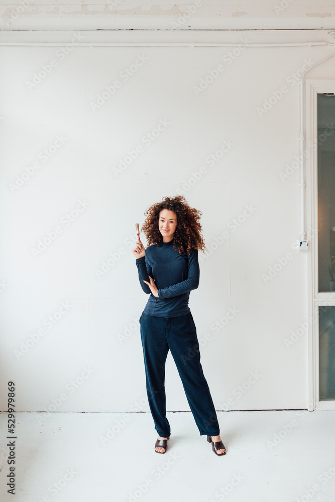 Beautiful woman with paintbrush standing in front of white wall Beautiful woman with paintbrush standing in front of white wall