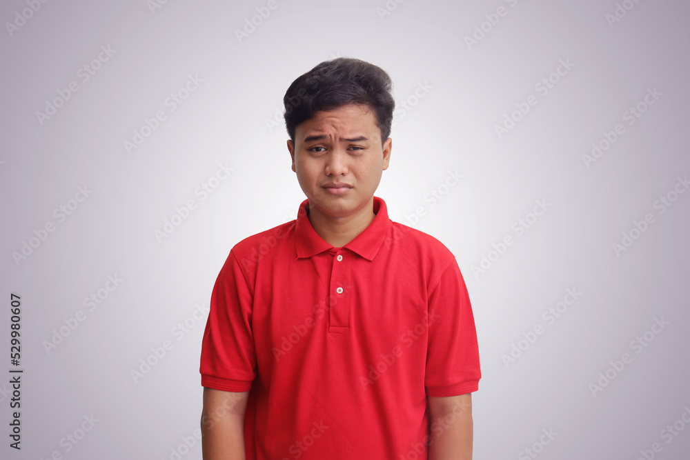 Portrait of unpleasant Asian man in red polo shirt showing his face sadness. Isolated image on gray background Portrait of unpleasant Asian man in red polo shirt showing his face sadness. Isolated image on gray background