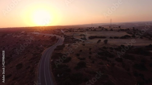 Aerial drone footage of a scenic rural street with a white pick up car passing leading to the sunset from above. Desert road with power cable lines, colourful sky and arid hills in Limassol, Cyprus. photo