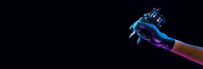 Closeup of tattooer master's hand in black glove holding machine for making tattoo art on body isolated on dark background in neon light.